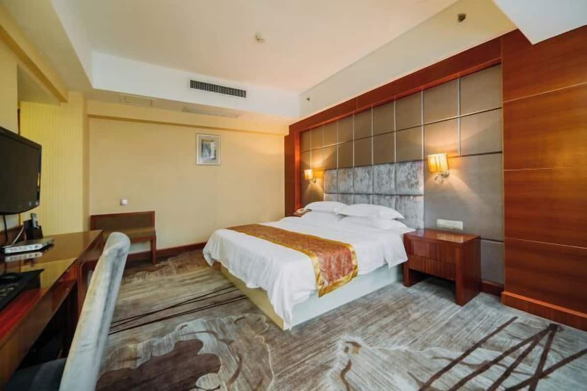 Quarto de hotel com uma cama grande, cabeceira de madeira, uma mesa com cadeira, televisão e uma mesinha.