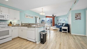 A modern kitchen with white cabinets, a granite countertop, and a built-in oven. There is a dining area with a table and chairs, and a living room with a blue sofa and a coffee table.