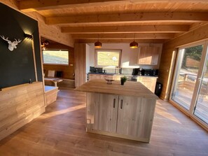 A spacious wooden cabin interior with a central island, large windows, and a mounted deer head on the wall.