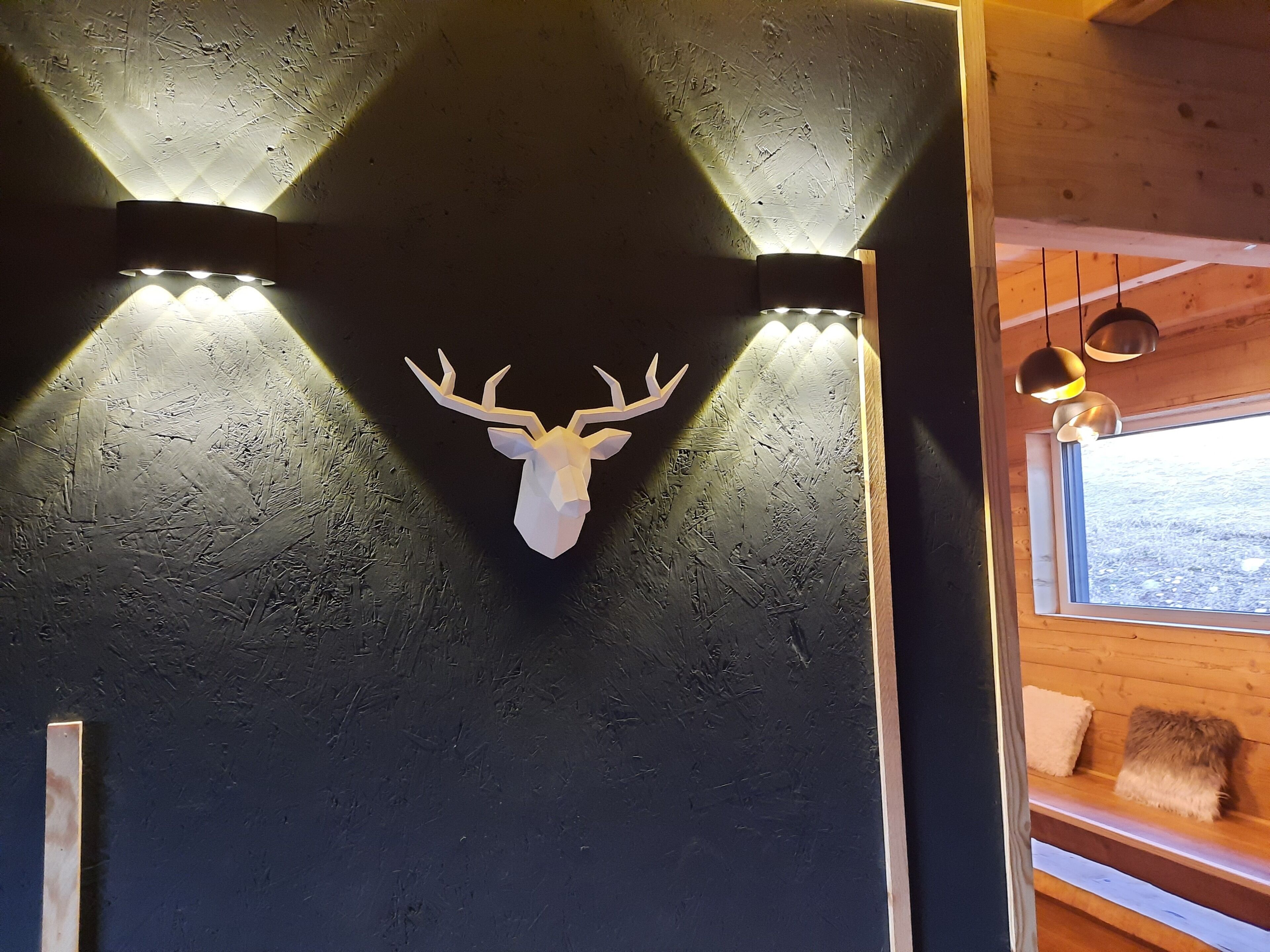 A wooden cabin interior with a mounted deer head on the wall, a window with a view of the outdoors, and a bench with a blanket.