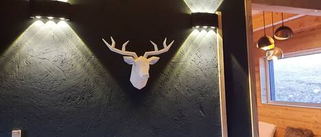 A wooden cabin interior with a mounted deer head on the wall, a window with a view of the outdoors, and a bench with a blanket.