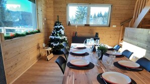 Une table de salle à manger en bois, mise pour six personnes, avec des assiettes, des verres et des couverts. Un sapin de Noël est visible à l’arrière-plan.