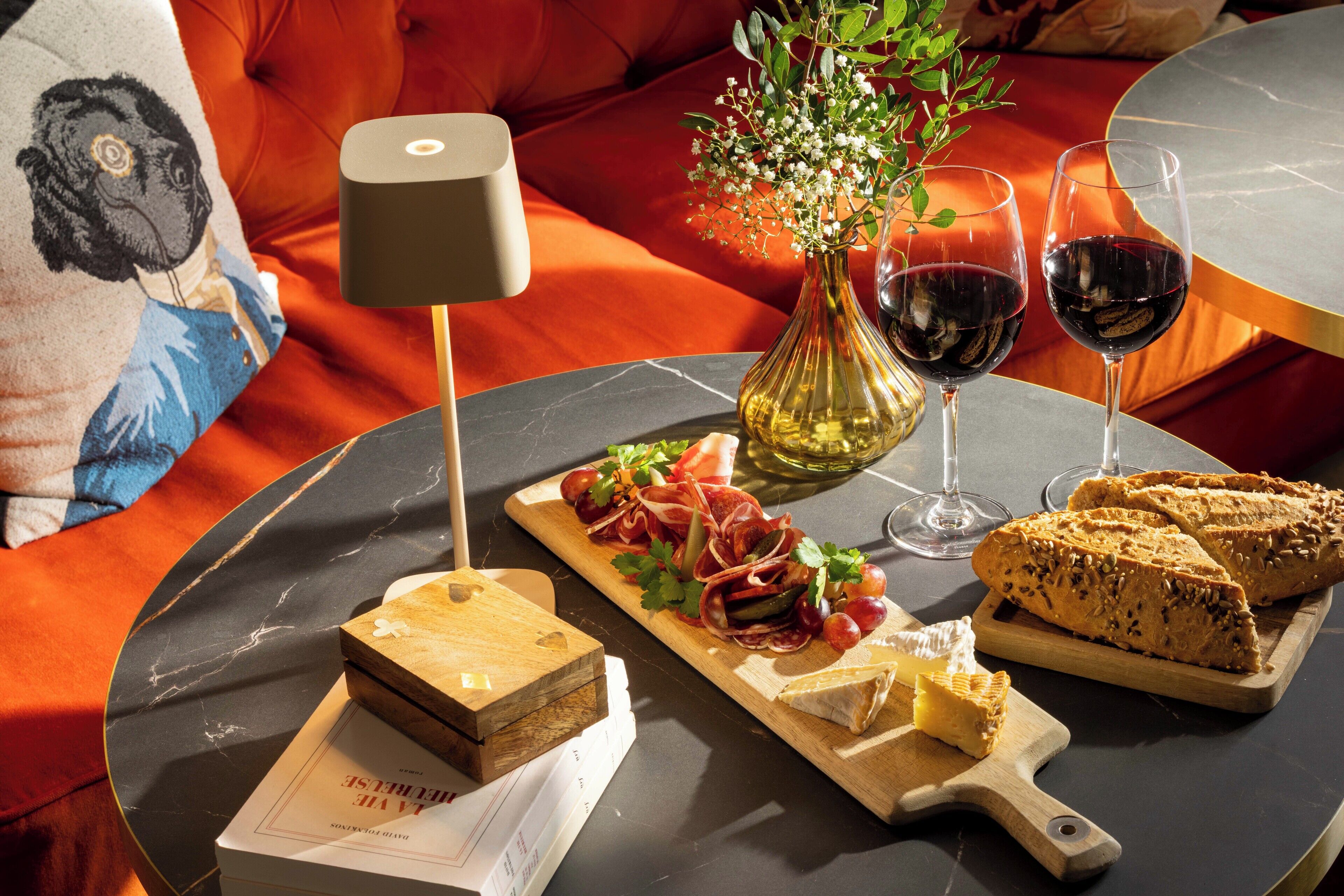 A dining table with a marble top, a vase with flowers, two glasses of red wine, a loaf of bread, a cheese board, and a small wooden box.