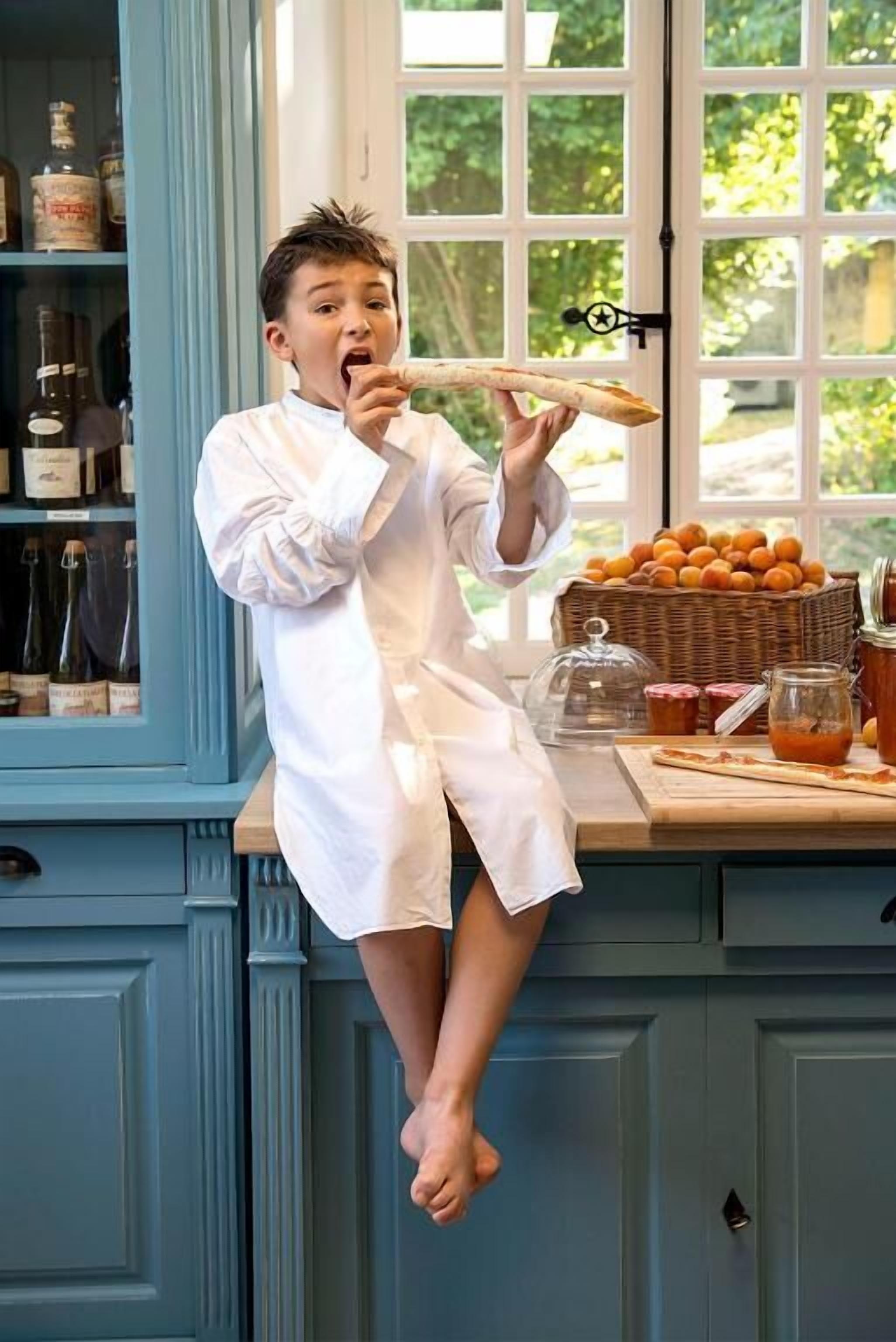 Una persona vestida con una bata blanca sentada en la encimera de una cocina, con una cesta de naranjas y frascos de conservas.