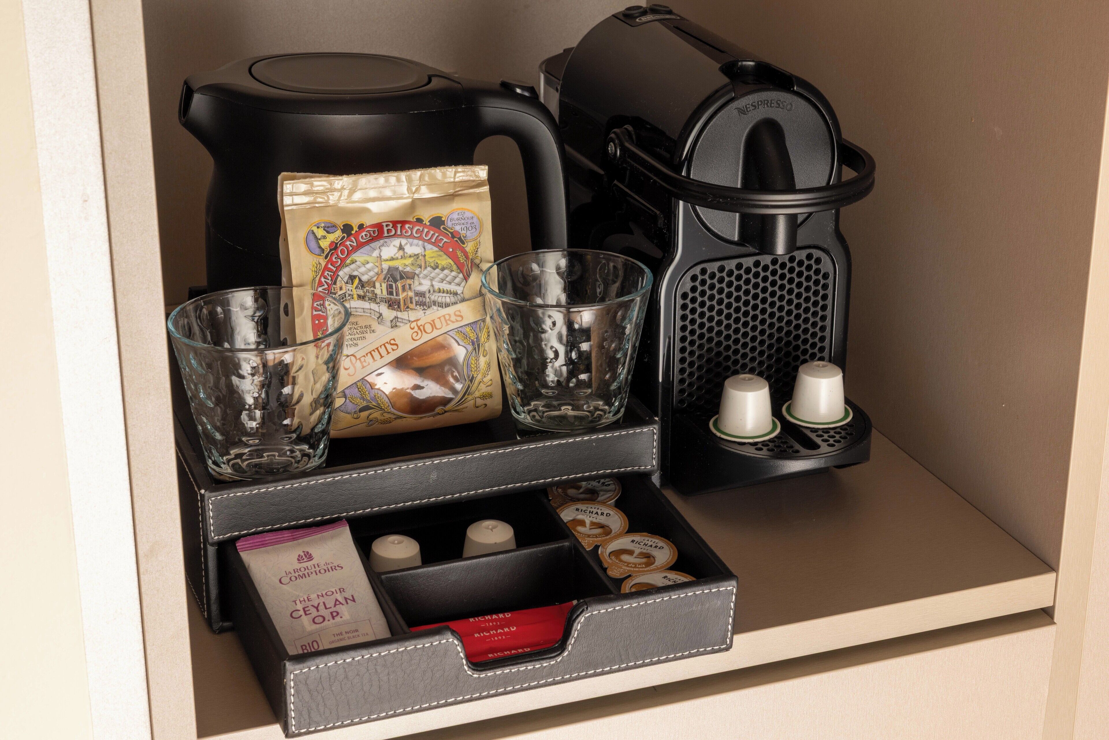 A coffee station with a coffee maker, a glass carafe, a bag of biscuits, and a compartment with coffee pods and a box of tea.