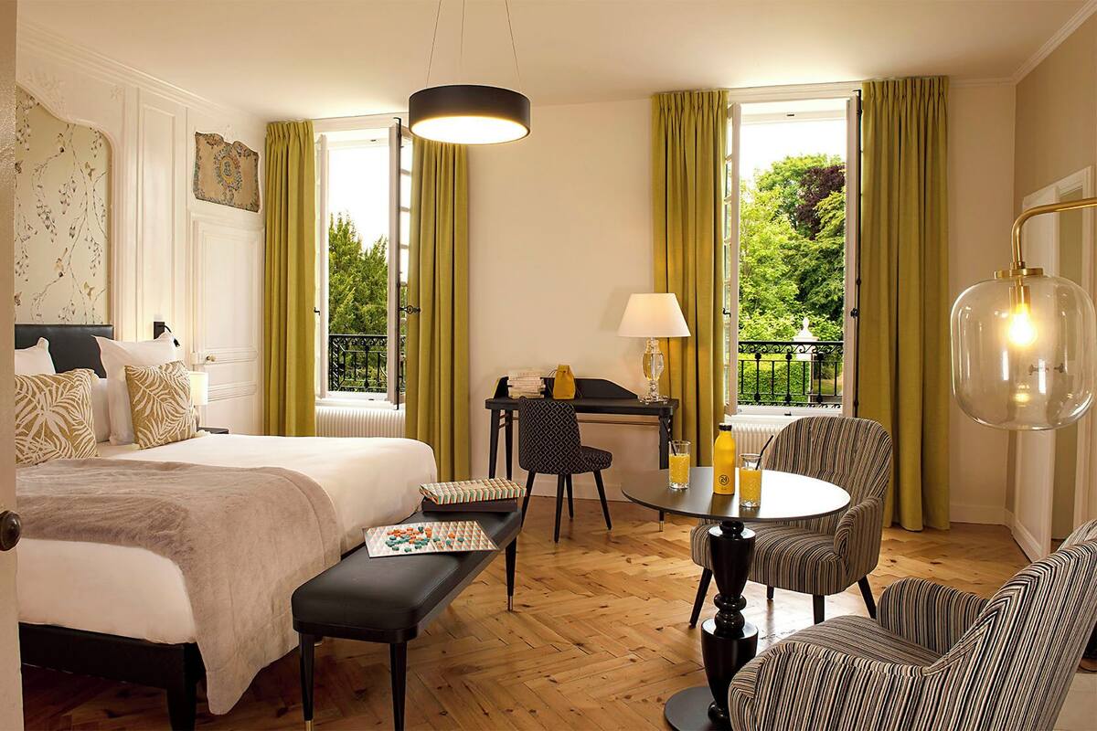 Une chambre d’hôtel avec un lit, un piano, une table à manger avec des chaises et une vue sur la verdure.