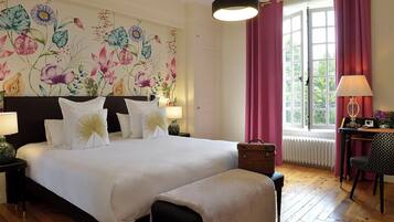 A hotel room with a large bed, a floral wall mural, a window with curtains, a table with a lamp, and a chair.