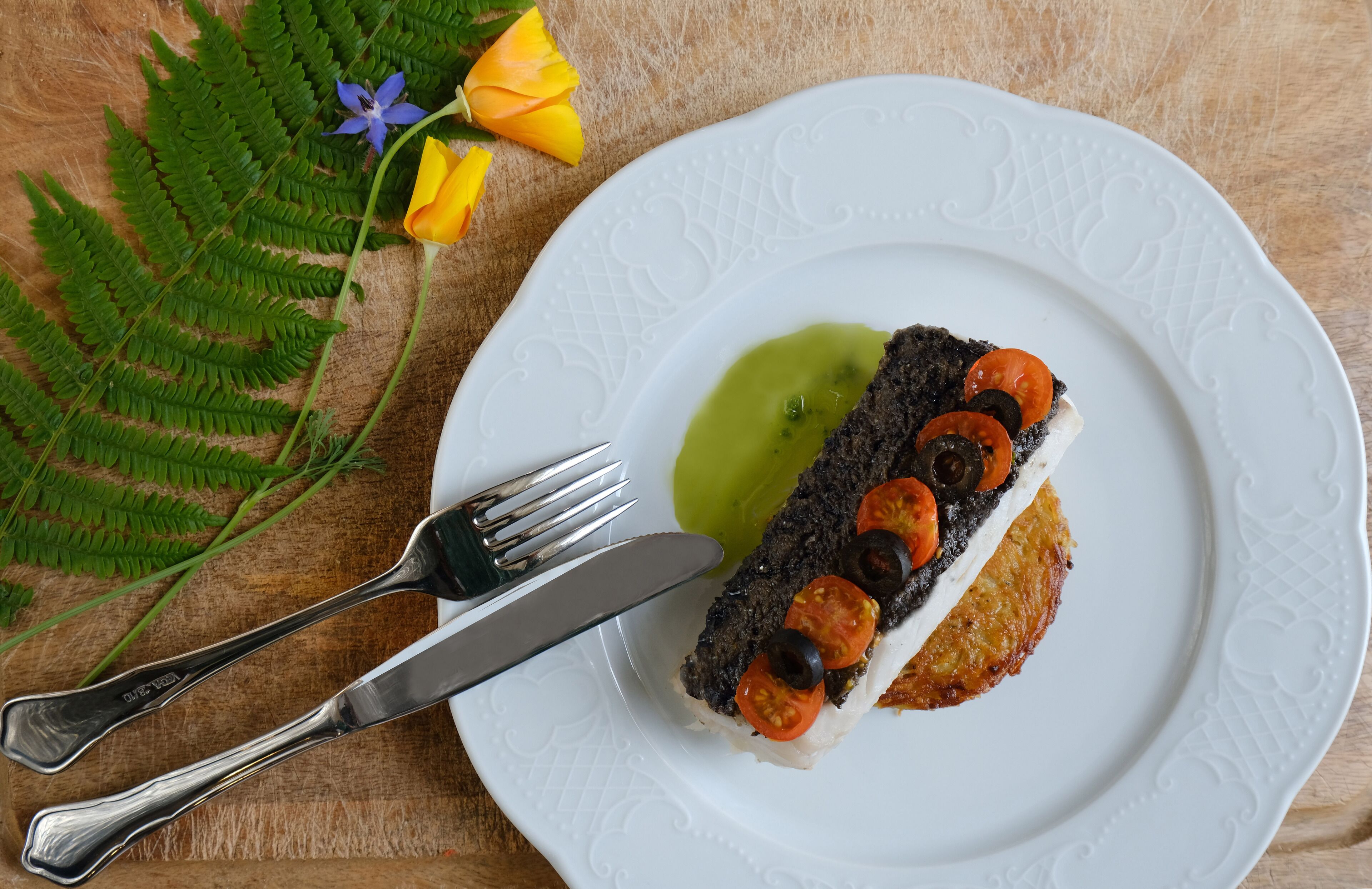 A white plate with a piece of food topped with black olives and cherry tomatoes, accompanied by a fork and knife.