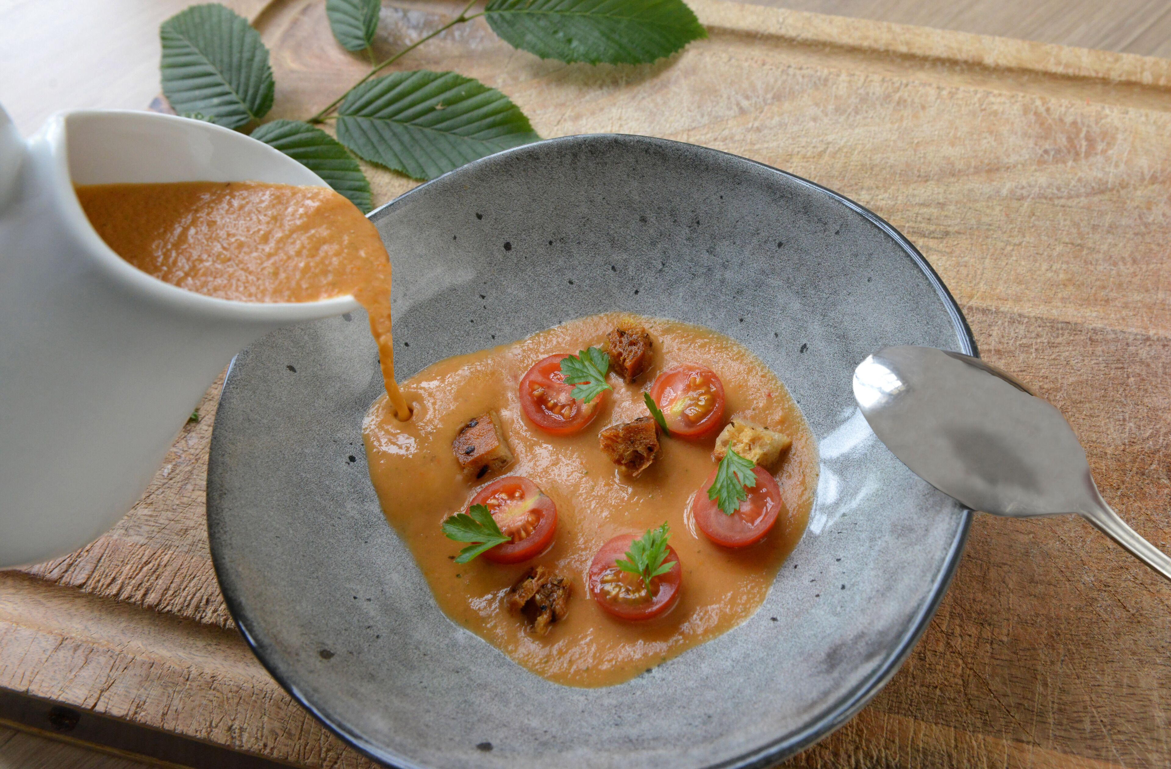 A bowl of soup with tomatoes and herbs on a wooden table.