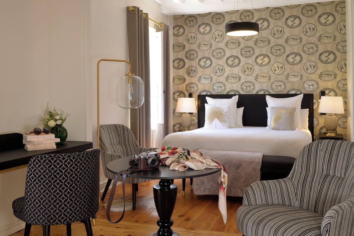 Une chambre d’hôtel avec un lit, deux chaises, une table sur laquelle se trouve un appareil photo, et un bureau avec des livres et un vase.