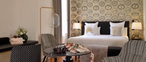 Une chambre d’hôtel avec un lit, deux chaises, une table sur laquelle se trouve un appareil photo, et un bureau avec des livres et un vase.