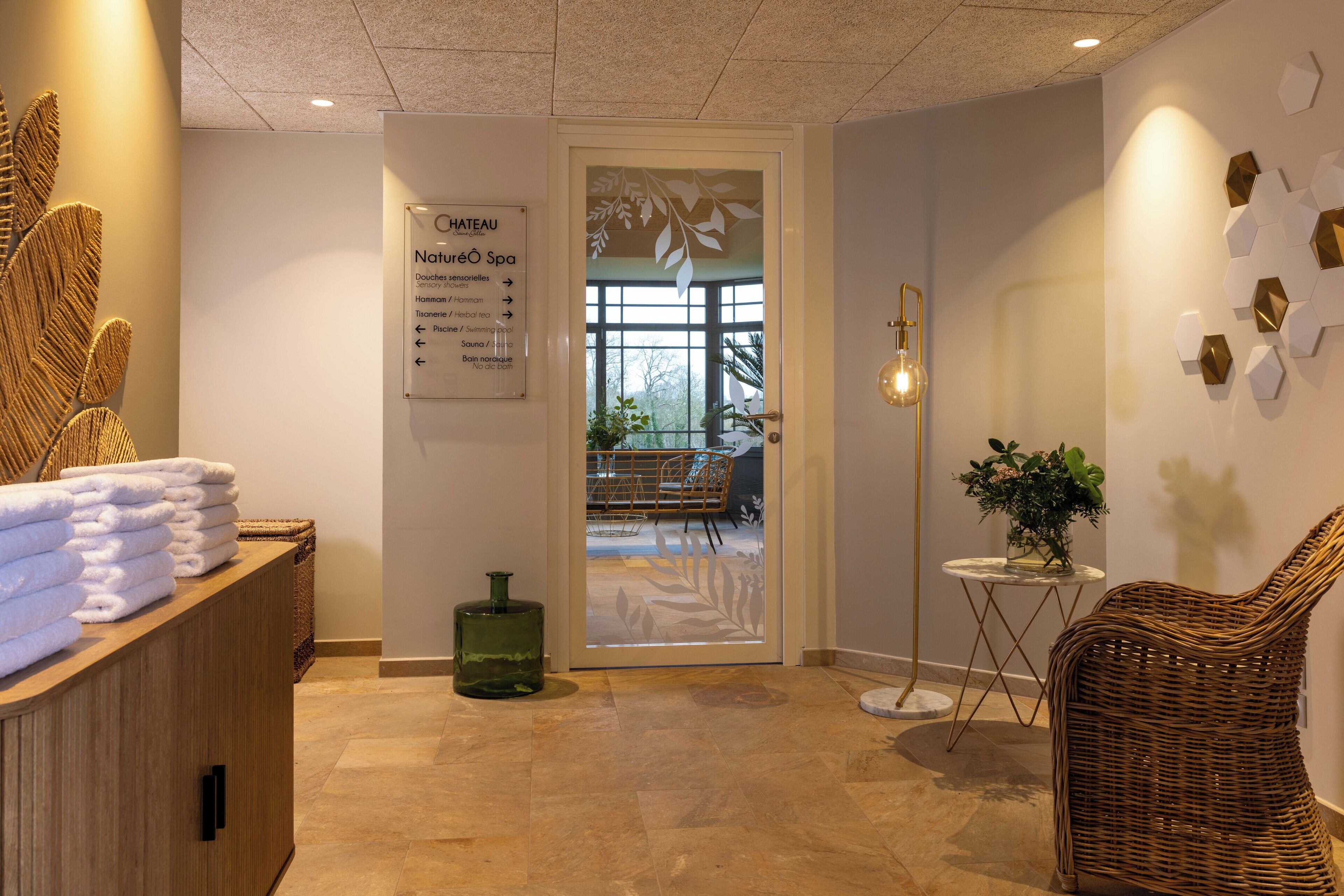 Un espace d’accueil de spa comprenant un panneau indicateur, un comptoir d’accueil avec des serviettes, un fauteuil en osier, une lampe sur pied et un mur orné de motifs hexagonaux.