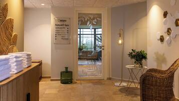 Un espace d’accueil de spa comprenant un panneau indicateur, un comptoir d’accueil avec des serviettes, un fauteuil en osier, une lampe sur pied et un mur orné de motifs hexagonaux.