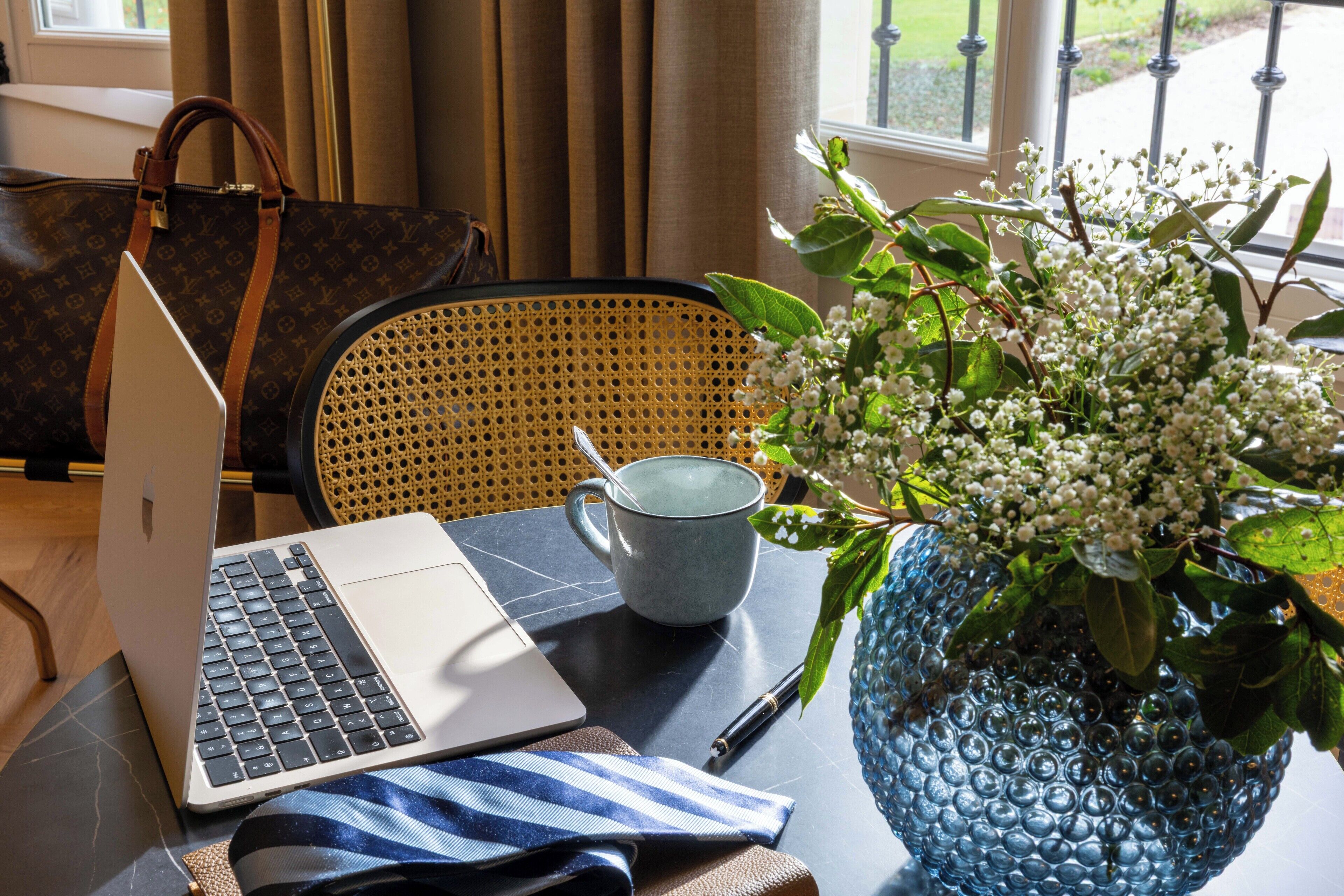Um laptop sobre uma mesa com um vaso azul de flores, uma caneca cinzenta e uma bolsa marrom.