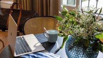 Um laptop sobre uma mesa com um vaso azul de flores, uma caneca cinzenta e uma bolsa marrom.