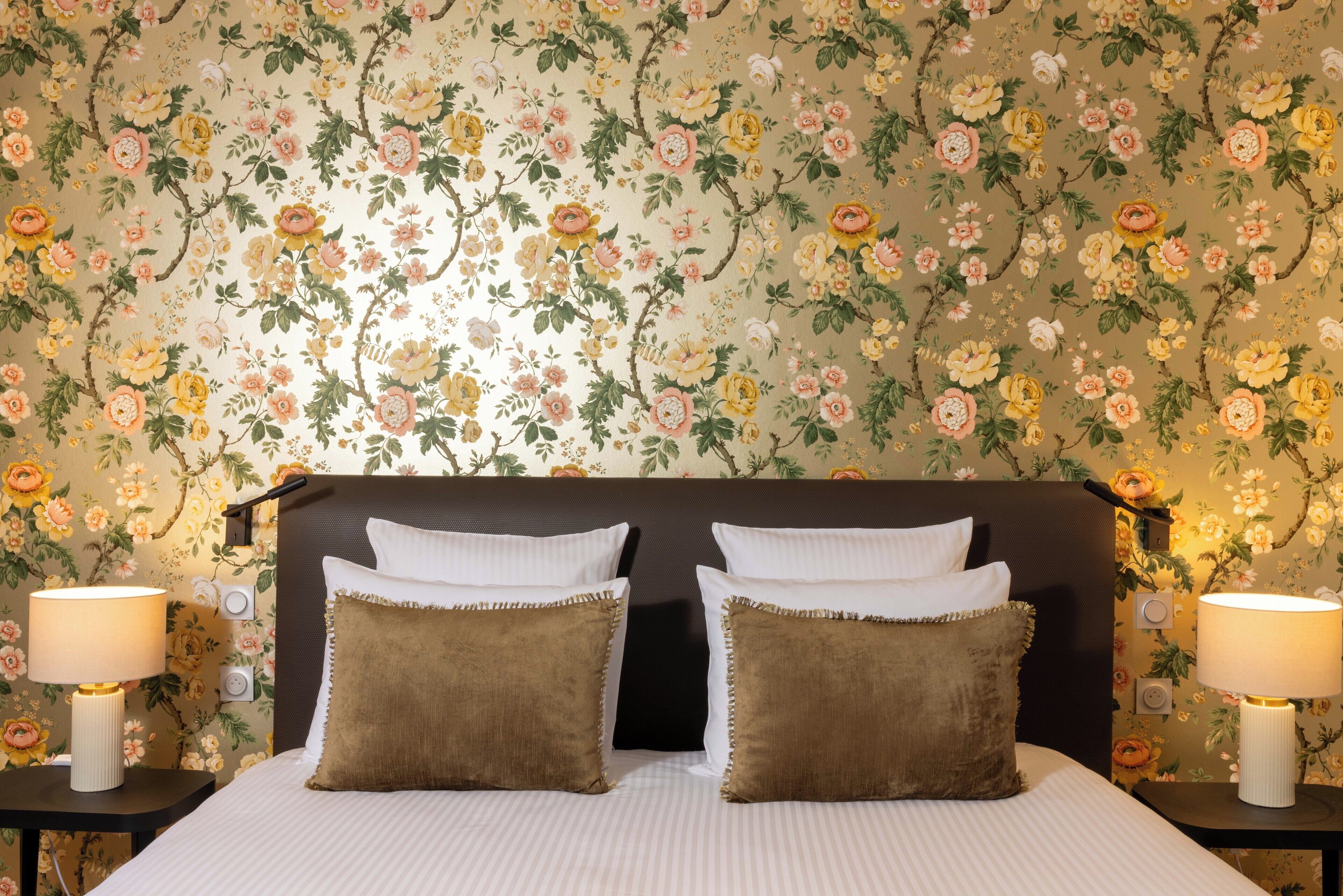 Un dormitorio con papel tapiz floral, una cama con almohadas blancas y marrones, y dos lámparas de noche.