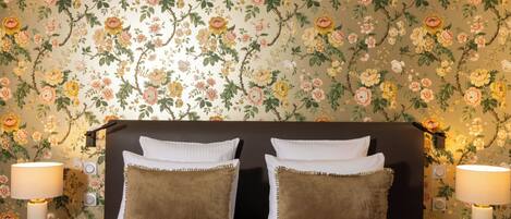 Un dormitorio con papel tapiz floral, una cama con almohadas blancas y marrones, y dos lámparas de noche.