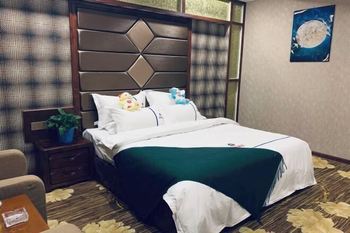 A neatly made bed with a green bedspread and white linens, a wooden headboard, a nightstand with a plant, and a wall with a framed artwork.