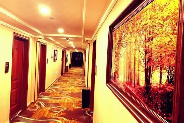 A hotel hallway with a patterned carpet, wooden doors, and a framed painting of a forest scene.