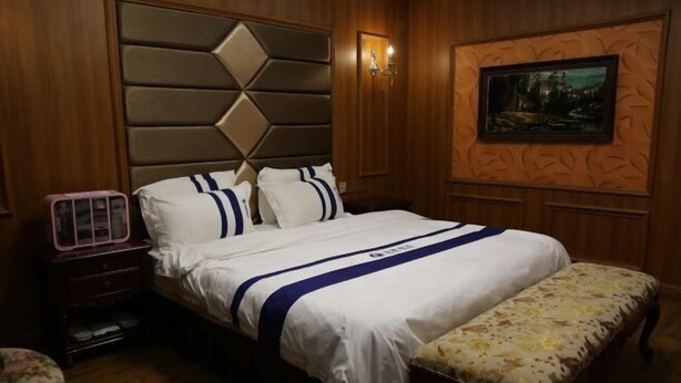 Un dormitorio con una cama, una mesita de noche, un banco, un cuadro colgado en la pared y una lámpara de pared.