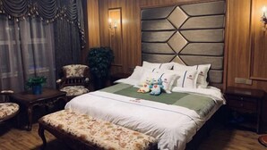 Habitación de hotel con cabecera de madera, cama con ropa blanca y almohadas mullidas, mesita de noche con lámpara, silla y pequeña mesa con una planta.