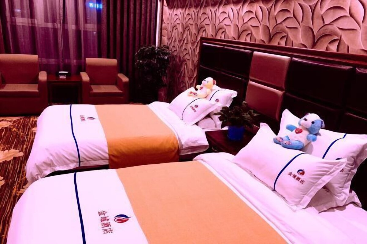 A hotel room with two beds, each with matching pillows and a logo on the bedsheet. There is a small table with a plant and a teddy bear between the beds.