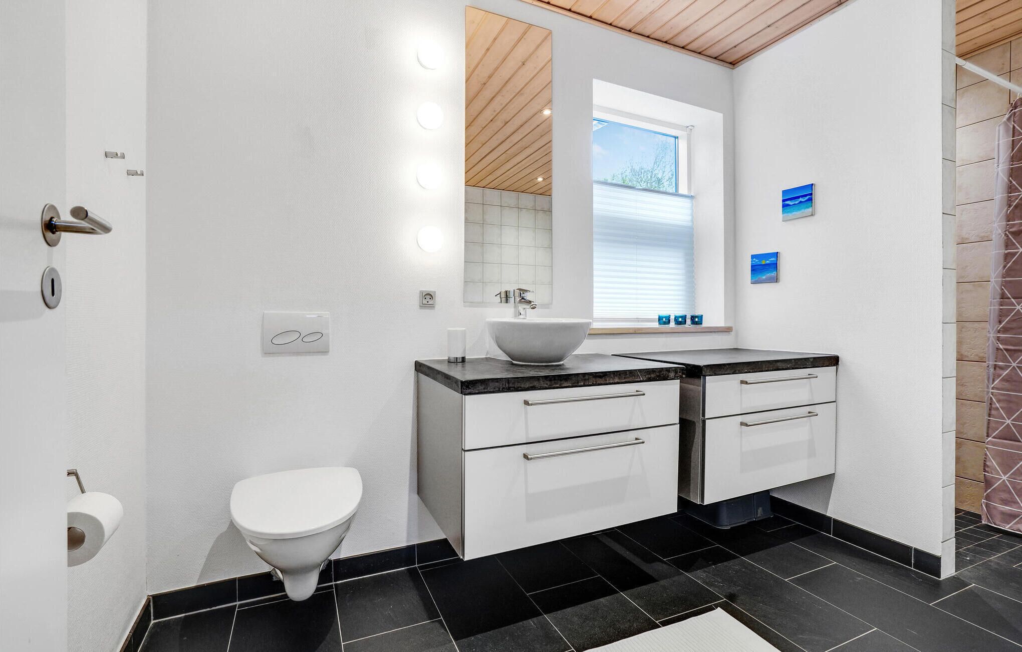 Une salle de bain moderne avec un lavabo blanc, deux tiroirs, des toilettes et une fenêtre avec des stores.