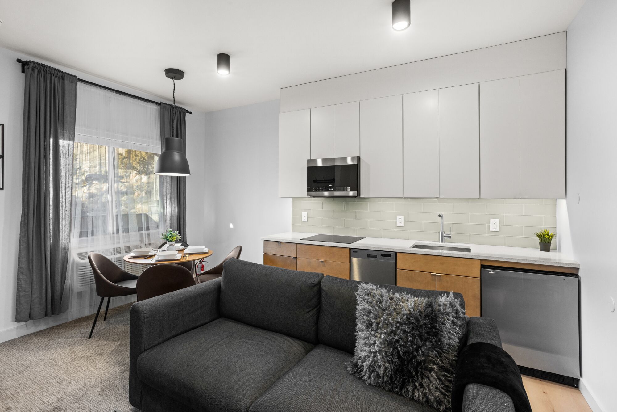 Een moderne keuken met grijze kastjes, een ingebouwde oven en een vaatwasser. Er is een eethoek met een tafel en stoelen, en een grijze bank met een zacht kussen.