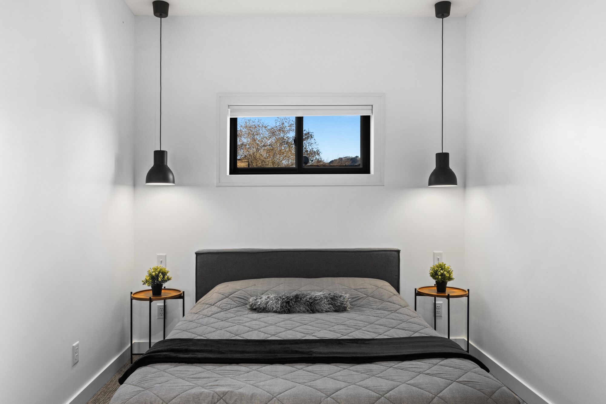 A bedroom with a bed, two bedside tables with plants, a window with a view of trees, and pendant lights.