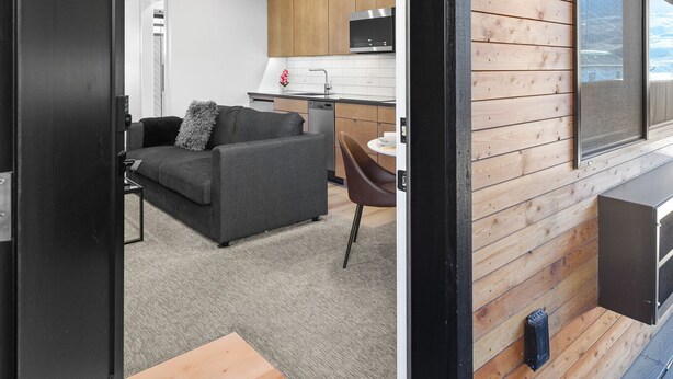 Eine offene Durchgangstür führt in einen Wohnbereich mit Sofa, Couchtisch und Esstisch. Im Hintergrund ist eine Küche mit Holzausstattung und einer Mikrowelle zu sehen.
