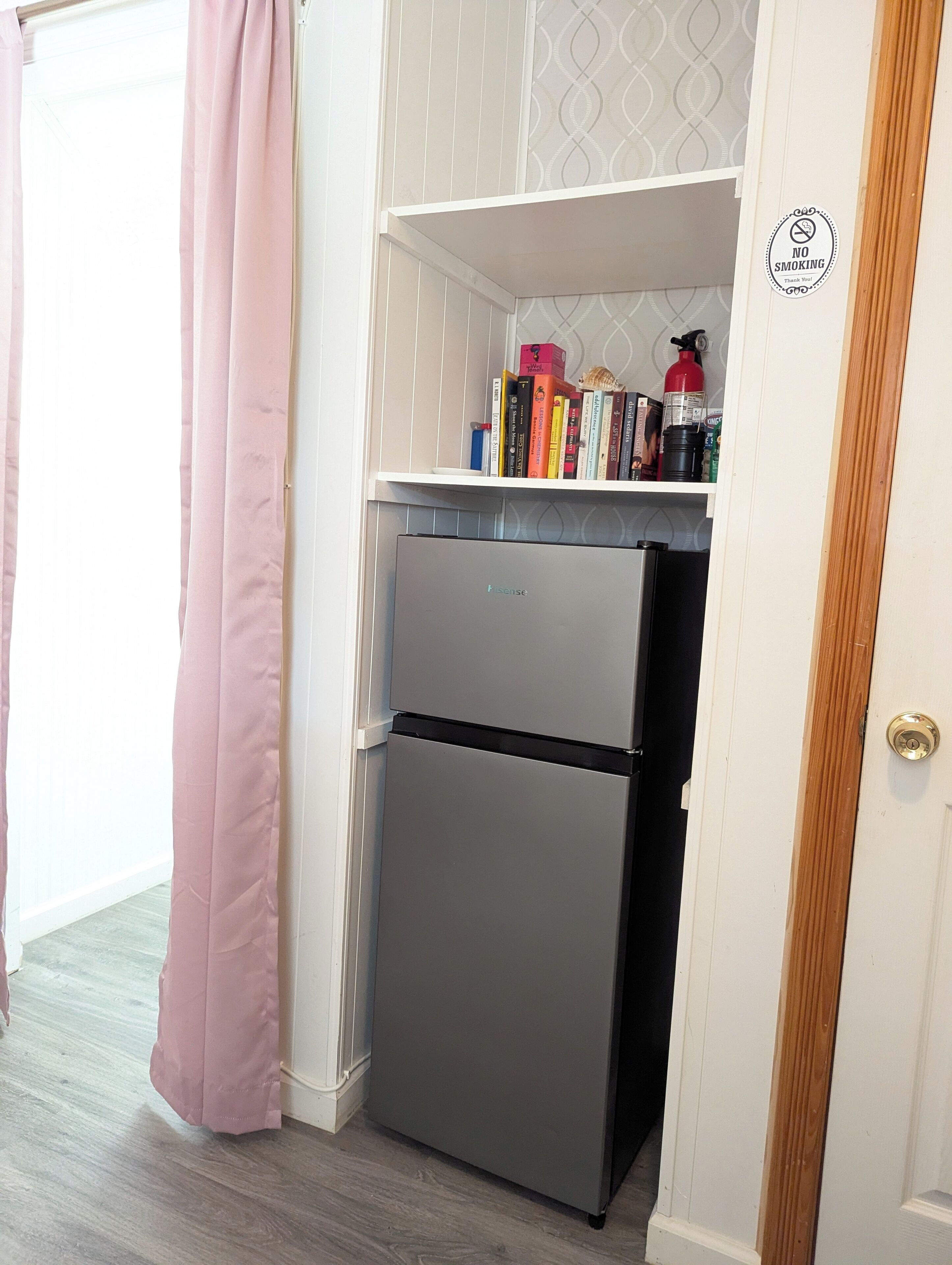 A kitchen with a grey refrigerator, a white shelf with books and a fire extinguisher, and a pink curtain.