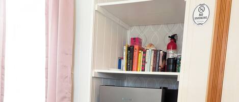 A kitchen with a grey refrigerator, a white shelf with books and a fire extinguisher, and a pink curtain.