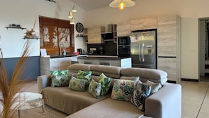 Een moderne woonkamer met een bank, een glazen salontafel en een keuken met een koelkast en fornuis.
