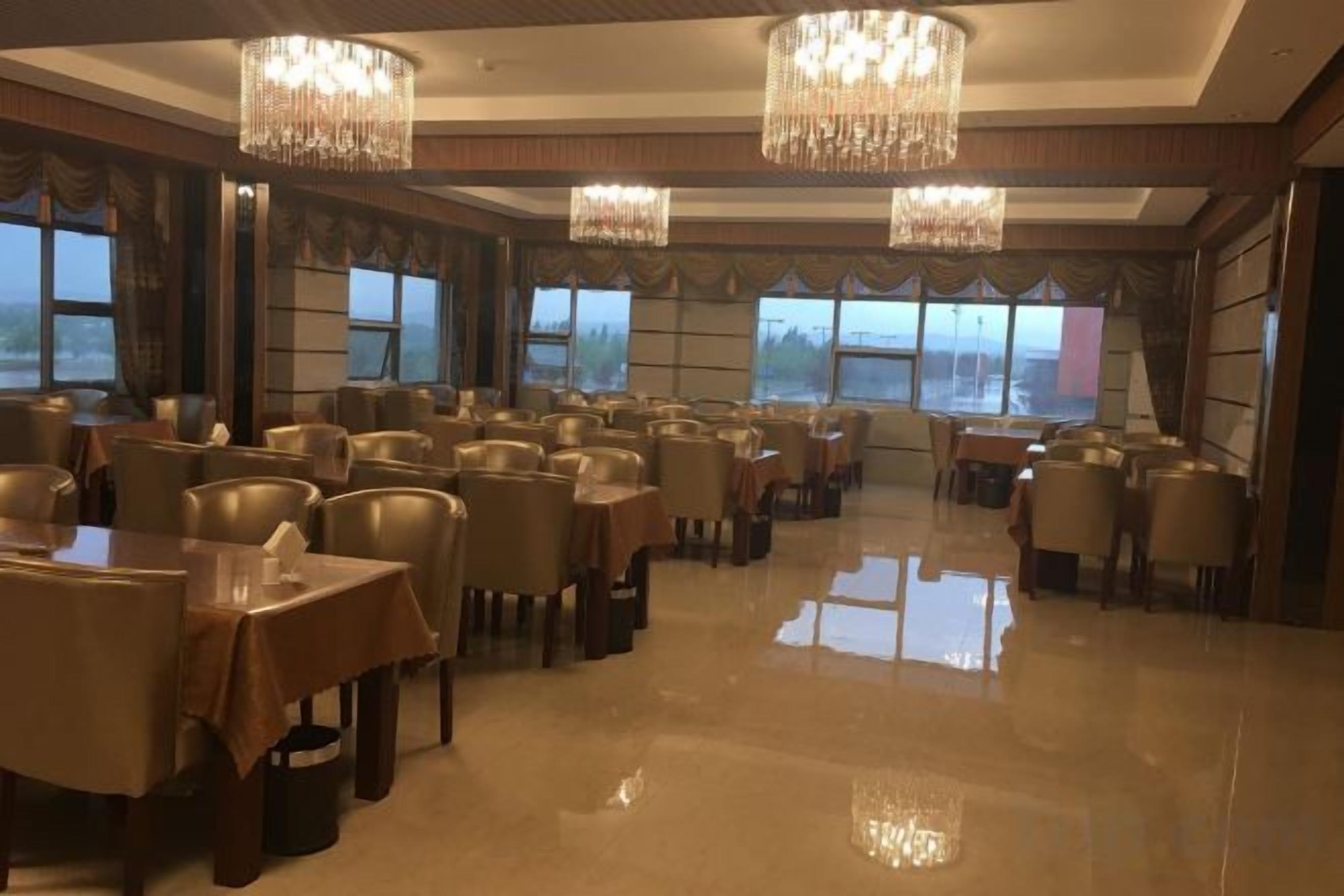 A hotel dining area with multiple tables and chairs, large windows, and chandeliers.