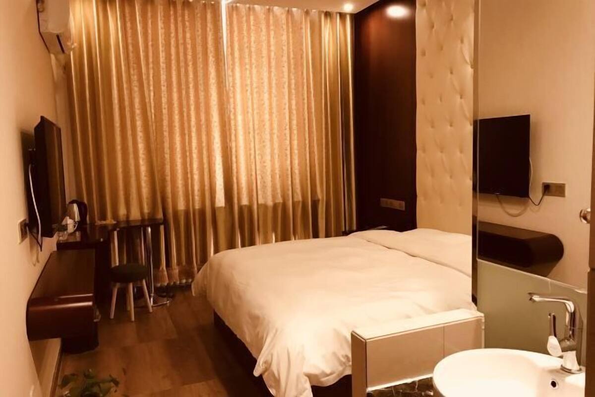 A hotel room with a bed, a desk, a chair, a TV, and a bathroom with a sink and mirror.