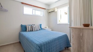 Un dormitorio con cama, mesita de noche, equipo de aire acondicionado, ventana con cortinas y un escritorio de madera.