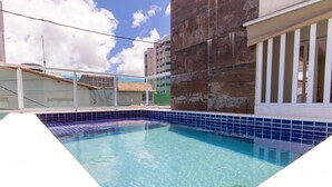Una piscina con agua cristalina azul, rodeada por una solana con baldosas y un edificio con balcón.