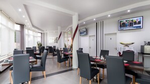 A modern dining area with multiple tables and chairs, a flat-screen TV mounted on the wall, and a large window providing natural light. - Turmhotel Rhein Main (Dreieich)