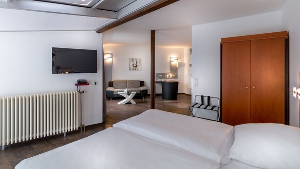 A modern hotel room with a large bed, a TV mounted on the wall, a radiator, a wooden door, and a small seating area with a sofa and table. - Turmhotel Rhein Main (Dreieich)