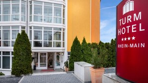 A modern building with a glass facade, a red sign for the Turm Hotel, and a paved entrance area with potted plants. - Turmhotel Rhein Main (Dreieich)