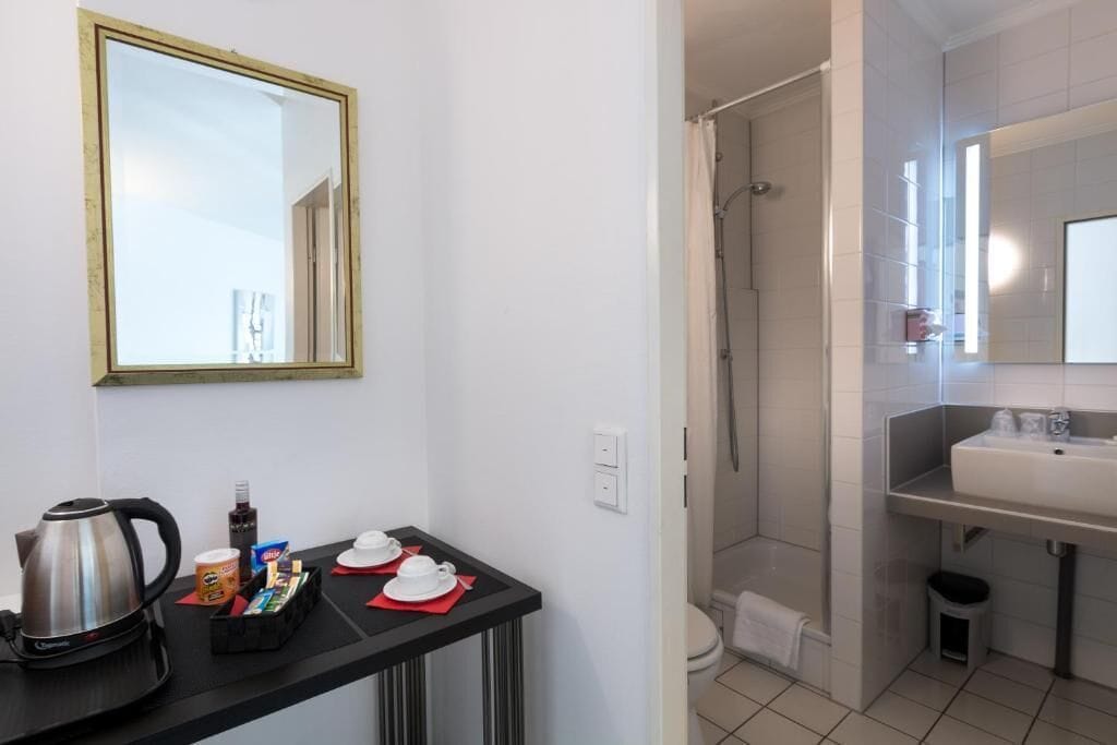 A hotel bathroom with a shower, sink, and mirror. There is a small table with a kettle, cups, and a bottle.