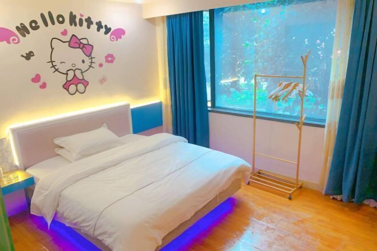 Een bed met witte lakens, een Hello Kitty muursticker, een raam met blauwe gordijnen en een houten rek.