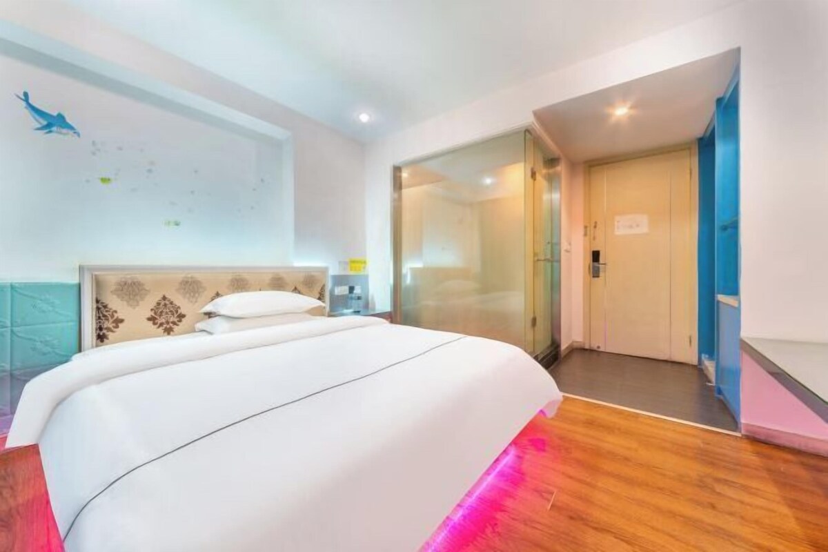 A hotel room with a bed, a nightstand, a wall-mounted clock, and a door leading to another room.