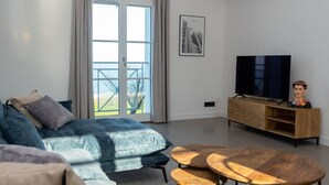 A modern living room with a sofa, a coffee table, a TV on a wooden stand, and a balcony with a view.