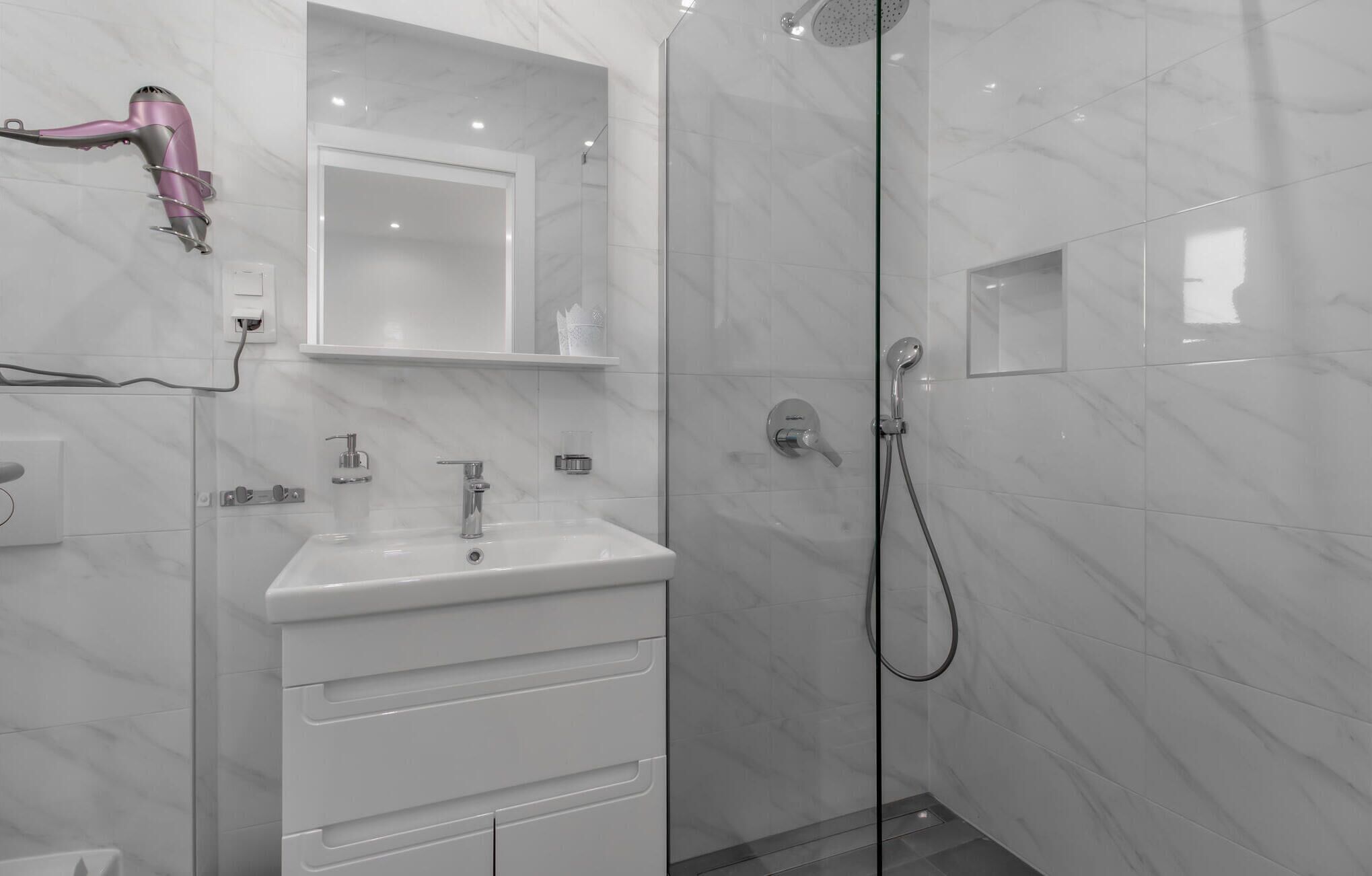 A modern bathroom with a marble shower, a sink with a mirror above it, and a hairdryer mounted on the wall.