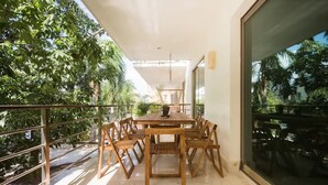 A wooden dining table with chairs on a balcony with glass railings, surrounded by lush greenery.