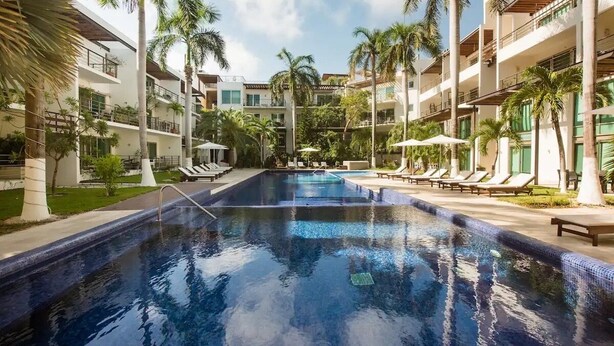 A swimming pool surrounded by palm trees and lounge chairs, with a multi-story building in the background.