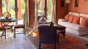 A cozy room with a white sofa, a wooden coffee table, and a chair. There is a view of a garden through large windows with curtains. - Small charming villa (Tamesluht)