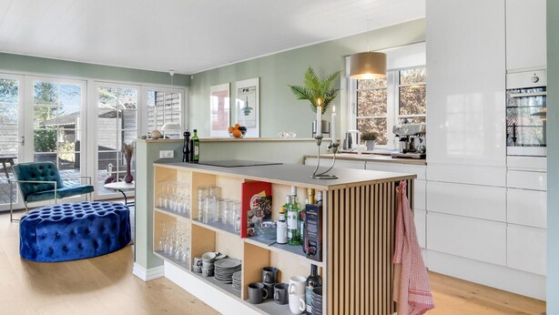 A modern kitchen with a central island, open shelving with glassware, and a seating area with a blue ottoman. - 4 bedroom lovely home in Vejby (Vejby)