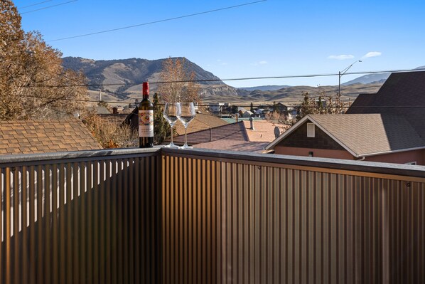 A balcony with a bottle of red wine and two glasses, overlooking a residential area with houses and mountains in the background.